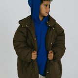 Child wearing an oversized brown puffer jacket open over a blue hoodie and white pants.