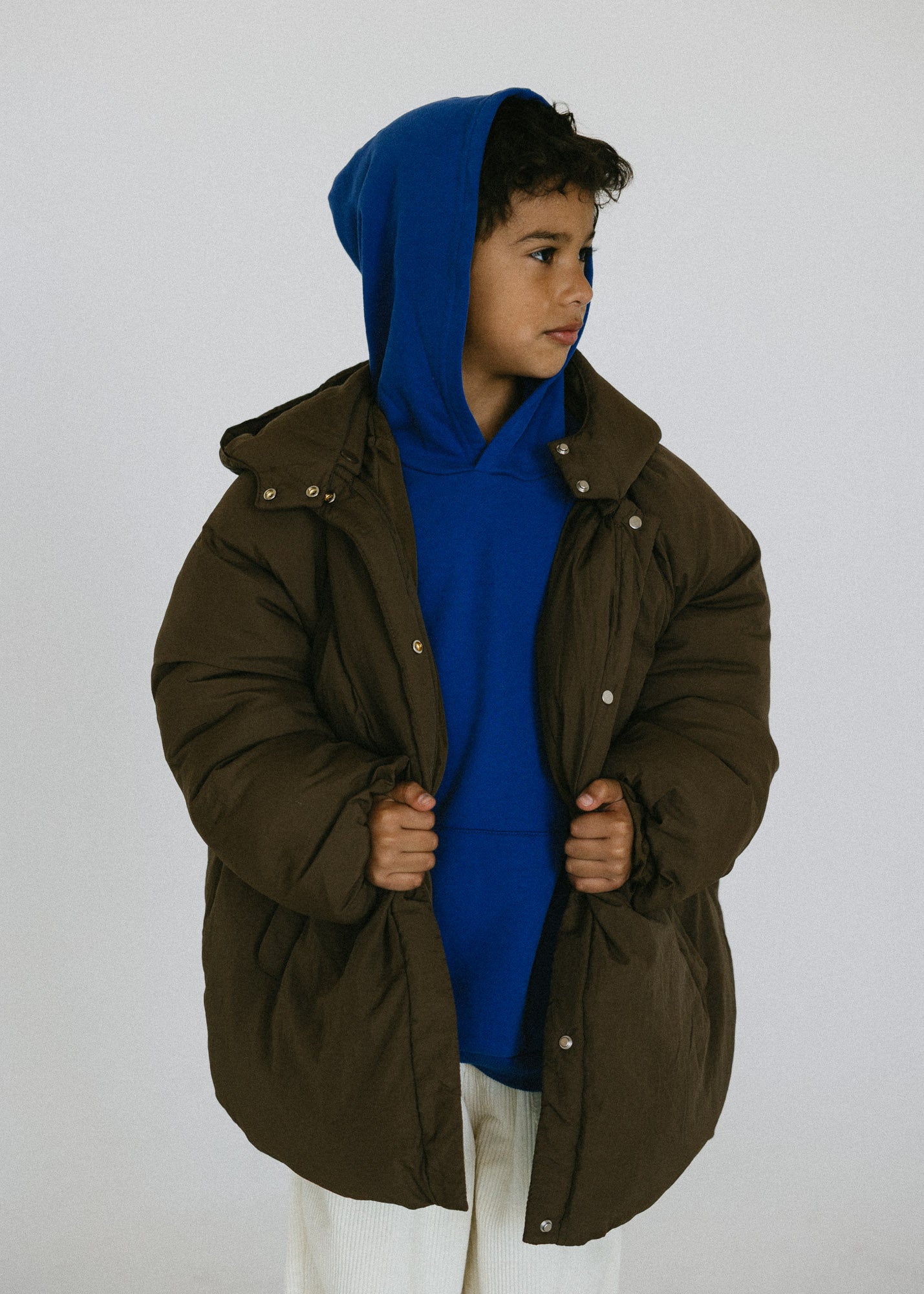 Child wearing an oversized brown puffer jacket open over a blue hoodie and white pants.