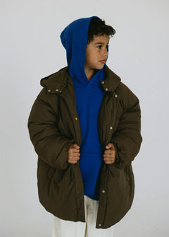 Child wearing an oversized brown puffer jacket open over a blue hoodie and white pants.