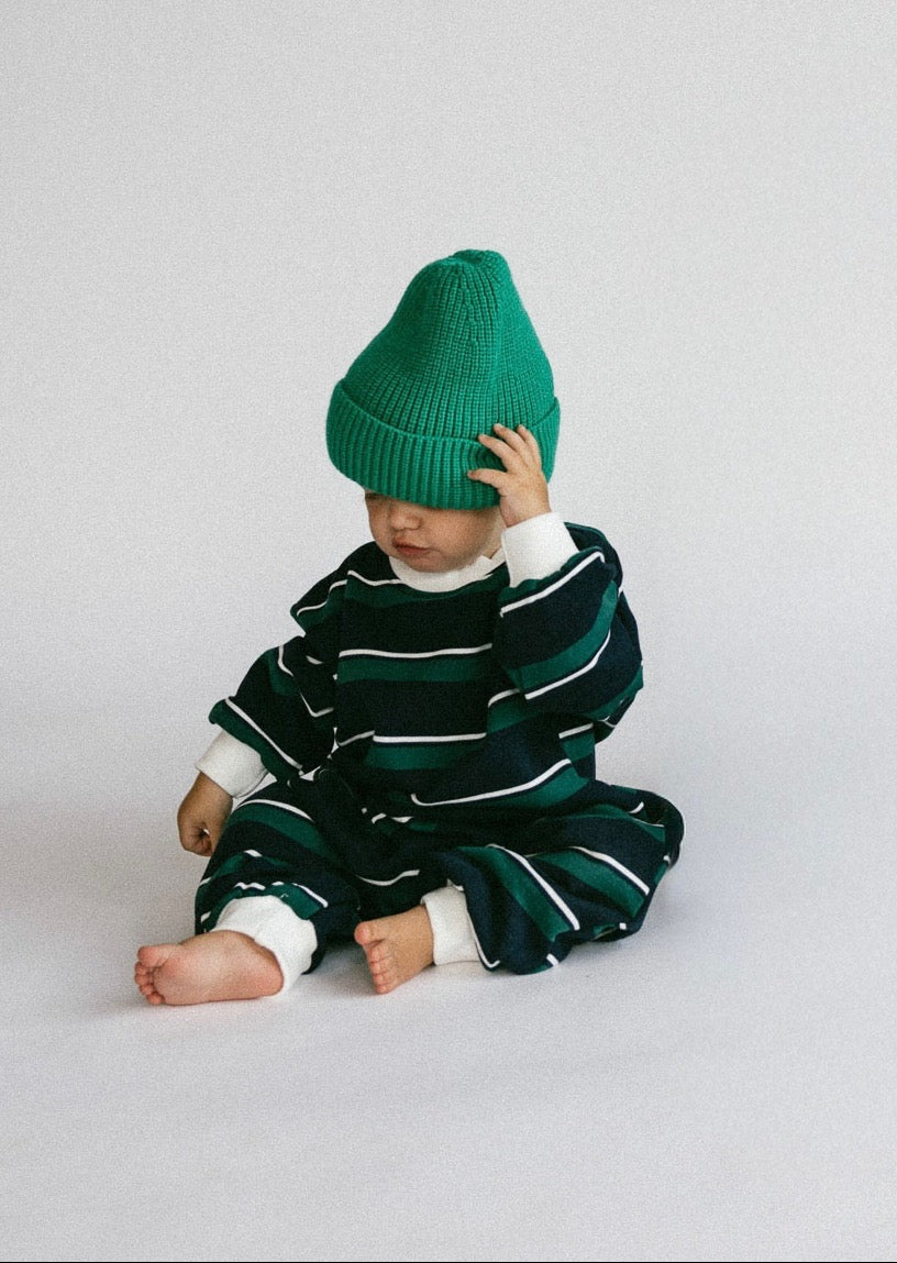 A baby sitting on a light gray studio floor, wearing a dark green and white striped knit romper and a bright green beanie hat.