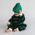 A baby sitting on a light gray studio floor, wearing a dark green and white striped knit romper and a bright green beanie hat.