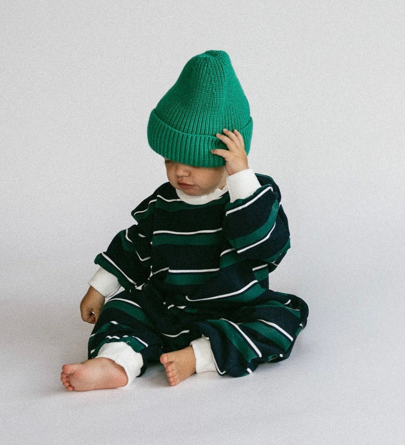 A baby sitting on a light gray studio floor, wearing a dark green and white striped knit romper and a bright green beanie hat.