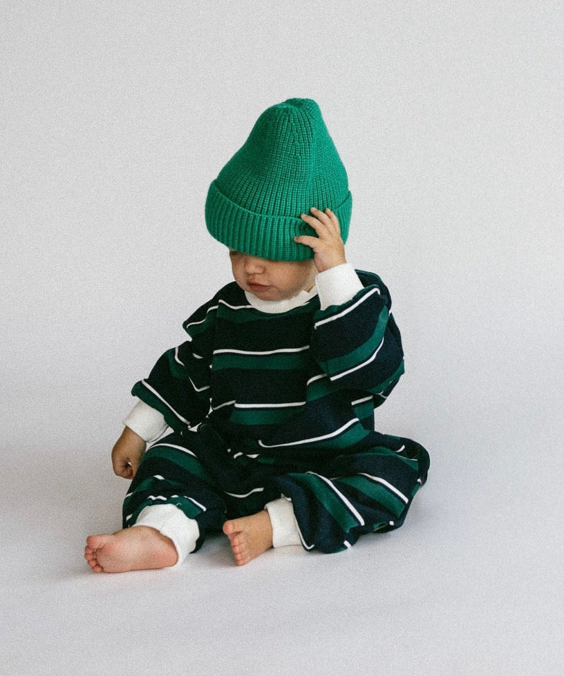 A baby sitting on a light gray studio floor, wearing a dark green and white striped knit romper and a bright green beanie hat.