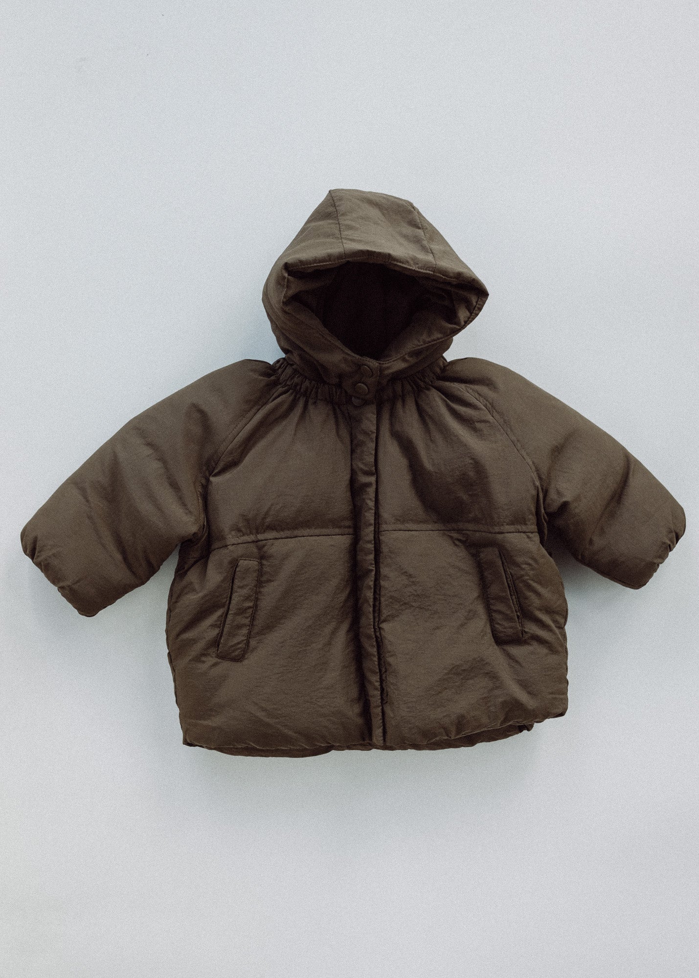 Front view of a brown kids puffer jacket laid flat, showing the hood, snap front and side pockets.