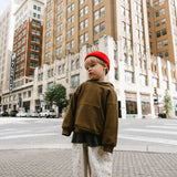 A child standing on a city street corner wearing an oversized brown hoodie, cream patterned pants, black sneakers, and a bright red knit beanie, with tall urban buildings in the background.