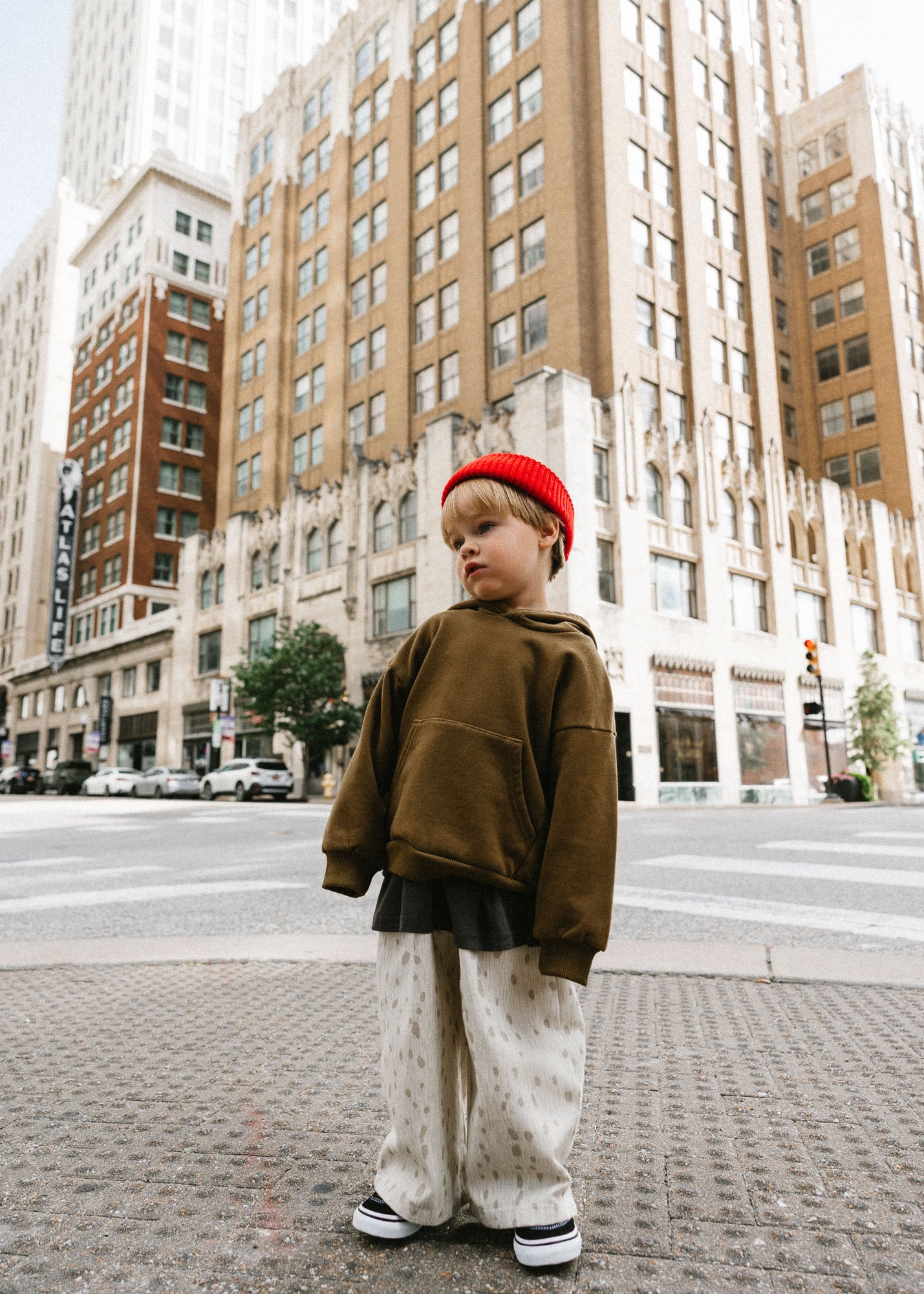 A child standing on a city street corner wearing an oversized brown hoodie, cream patterned pants, black sneakers, and a bright red knit beanie, with tall urban buildings in the background.