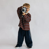 A young boy with blond hair stands against a light gray studio background, holding a vintage-style camera to his eye. He is wearing a long-sleeve striped shirt in red, brown, and white, dark navy wide-leg pants with white side stripes, and black sneakers.