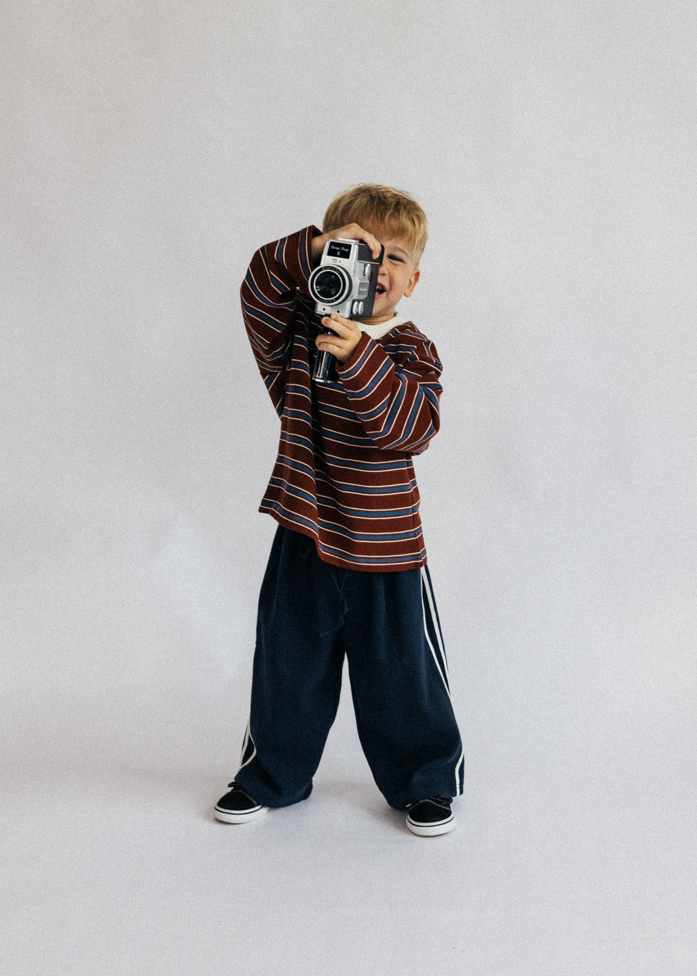 A young boy with blond hair stands against a light gray studio background, holding a vintage-style camera to his eye. He is wearing a long-sleeve striped shirt in red, brown, and white, dark navy wide-leg pants with white side stripes, and black sneakers.